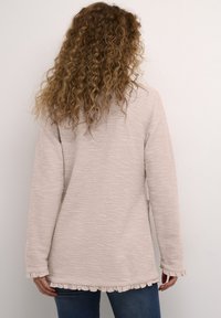 Jasnoróżowa, teksturowana bluzka z długim rękawem, z falbankami na dole i mankietach, o luźnym kroju i miękkim materiale, noszona ze spodniami jeansowymi w kolorze niebieskim.