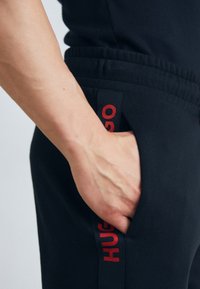 Črne trenirke imajo stransko žep z rdečim logotipom "HUGO". Pas je elastičen, kar zagotavlja udobno prileganje. Gladka struktura tkanine.