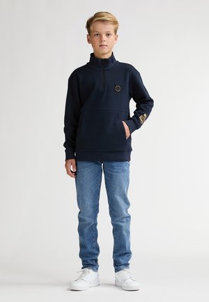 Marineblauwe hoodie met rits en hoge kraag, voorvak en geborduurd logo; gecombineerd met lichtblauwe spijkerbroek en witte sneakers.