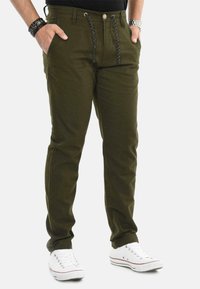 Man wearing olive green casual pants with black drawstrings, black shirt, white sneakers, black bracelet on left wrist, and silver watch on right wrist.