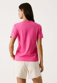 Fuchsia Baumwoll-T-Shirt mit kurzen Ärmeln, rundem Halsausschnitt und dezenter Markenkennzeichnung auf der Rückseite. Kombiniert mit hellbeigen, ausgefransten Shorts.
