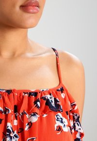 Gros plan sur la partie inférieure du visage, le cou et l'épaule d'une femme portant un haut à bretelles fines rouge à motifs floraux avec un détail à volants.