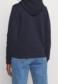 Granatowa bluza z kapturem na sznurek, z prążkowanymi mankietami i dolnym pasem. Połączona z jasnymi jeansami w jasnym odcieniu z przeszyciami na tylnej kieszeni.