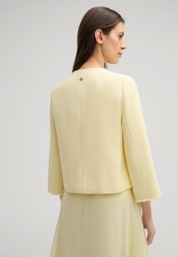 Giacca corta a maniche corte in tessuto giallo pallido testurizzato, con scollatura tonda e dettagli di bottoni sul retro.