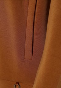 Bruine stof met een gladde textuur en een verticale zakdetail. Bevat een kleine metalen accent nabij de zoom voor functionaliteit.
