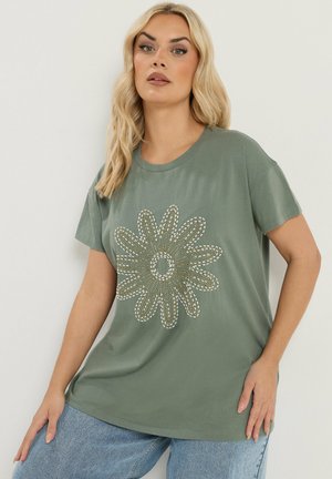 Donna con capelli biondi che indossa una maglietta verde con un grande motivo floreale bianco e oro, abbinata a jeans azzurri.