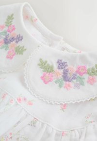 Bela bluza z okroglim ovratnikom, okrašena z roza, vijoličnim in zelenim cvetličnim vezenjem, čipkasto obrobo in mehkobo teksturirano tkanino.