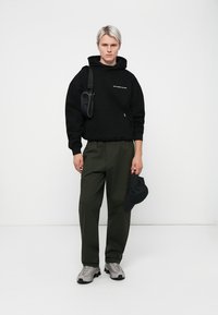 Zwarte hoodie met de tekst "VERSPIL GEEN KLEDING", olijfgroene cargo pants, grijze sneakers en een zwarte pet in de hand.