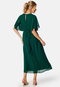 Zelené šifónové maxi šaty s jemnými rukávy, klíčovým otvorem na zádech a uvázaným pasem. Průsvitná textura s volným siluetou. Čisté barevné akcenty.