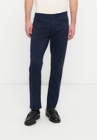 Chinos en coton bleu marine à coupe slim, avec fermeture à bouton sur le devant et poches latérales, assortis à des chaussures en cuir noir.