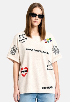 Donna che indossa una maglietta beige oversize con testo e simboli neri e rossi, inclusi "I believe in the power of love" e "Karma" cuore, abbinata a occhiali da sole.