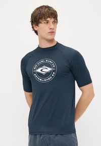 Mörkblå kortärmad surftröja tillverkad av lätt, stretchy material; har en cirkulär vit logotyp på bröstet med text och grafik.