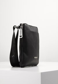 Svart läder crossbody-väska med texturerad finish, dragkedjestängning och ett märkesband. Har en subtil metallisk logotyp på framsidan.