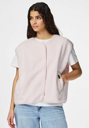 Femme aux longs cheveux foncés portant une veste en polaire rose clair boutonnée sur un t-shirt blanc et un jean bleu clair, une main dans la poche de la veste.