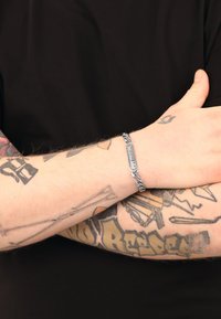 Pulsera de cadena de plata con placa rectangular grabada, con superficie lisa y eslabones entrelazados, llevada en un antebrazo con tatuajes.