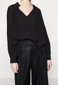 Femme portant un chemisier noir à manches longues avec un col en V et un pantalon noir plissé taille haute, sur un fond uni.