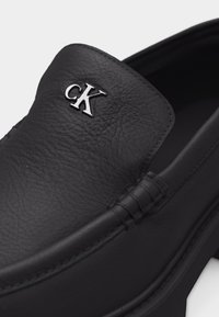 Chaussure à enfiler en cuir noir avec dessus texturé et logo argenté "cK" sur le cou-de-pied, avec détails cousus le long des bords.