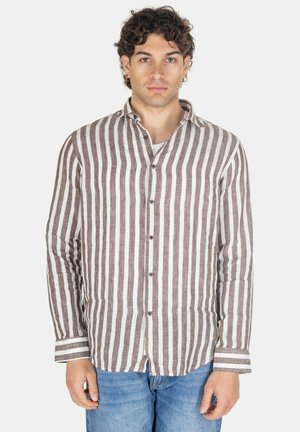 Uomo che guarda frontale con indosso una camicia a maniche lunghe a righe verticali marroni e bianche e jeans blu su uno sfondo semplice.