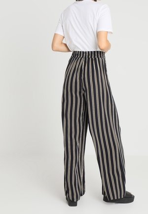Zwart en beige gestreepte wijde broek met een elastische tailleband. Soepele stoftextuur. Draagt een eenvoudig wit T-shirt en zwarte schoenen.