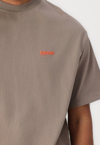 T-shirt in cotone grigio con maniche corte. Presenta un piccolo logo arancione "bareen" nell'area del petto. Tessuto morbido, design classico con collo a giro.