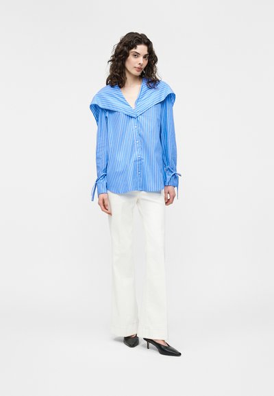 Γυναίκα που φοράει μπλούζα με μπλε και λευκές ρίγες και φαρδύ γιακά, λευκό παντελόνι φαρδιάς γραμμής και μαύρες γόβες με μυτερή μύτη, στέκεται μπροστά σε ένα απλό φόντο.