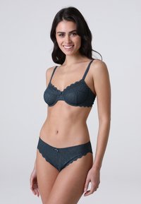 Set di lingerie in pizzo verde scuro con bordi scallop, reggiseno con ferretto e slip abbinati con un fiocco decorativo. Motivo geometrico testurizzato.