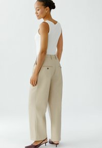 Beige taillierte Hose mit hoher Taille, zwei Gesäßtaschen und flachem Vorderteil. Ein enganliegendes, weißes Ripptanktop vervollständigt das Outfit.