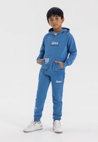 Blå hoodie och matchande joggers i mjukt material, med en framficka, grafisk text och rutigt detalj på fickan. Vita sneakers.