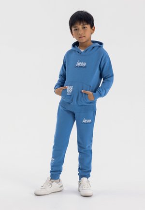 Blaues Kapuzenshirt und passende Jogginghose aus weichem Stoff, mit einer Fronttasche, grafischem Text und karierter Verzierung an der Tasche. Weiße Turnschuhe.