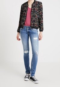 Blouson aviateur à motif floral en noir, chemise col en V rose, jean bleu déchiré, et baskets noires avec des accents blancs.