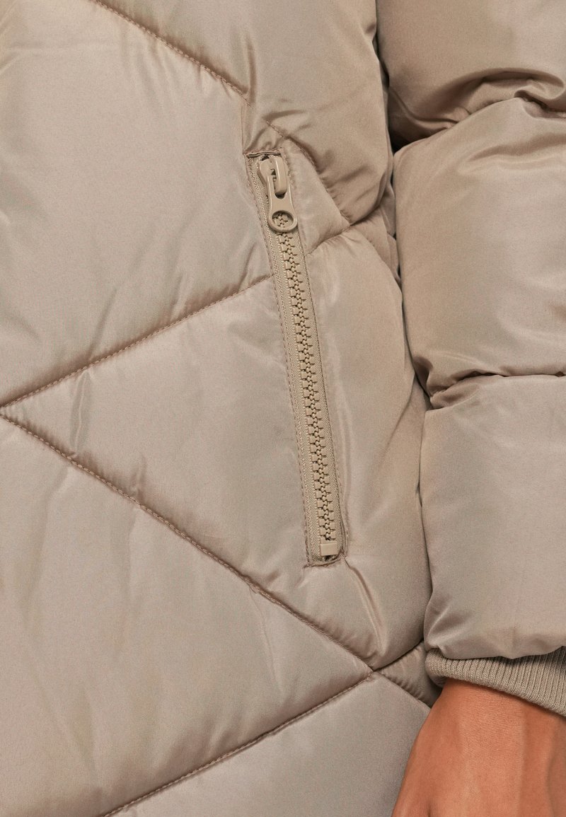 Gros plan sur une veste beige matelassée avec des coutures diagonales, une poche zippée beige et une manche à revers sur le bras d'une personne.