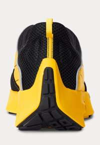 Svart och gul sportskor med en ovandel i mesh, en upphöjd häl och gummiyttersula med ett texturerat greppmönster. Gul dragflik baktill.