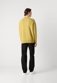 Overdimensjonert gul ullgenser med ribbet kant og mansjetter, sammen med svarte bukser og hvite joggesko, sett fra baksiden.