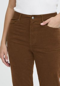 Brown corduroy pants with a slim fit, featuring a silver button, front pockets, and vertical ribbed texture. Light stitching detail visible.