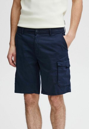 Férfi sötétkék halásznadrágban, oldalzsebes résszel és fehér ujjatlan felsőben, kezét a zsebében tartva, sima háttér előtt állva.