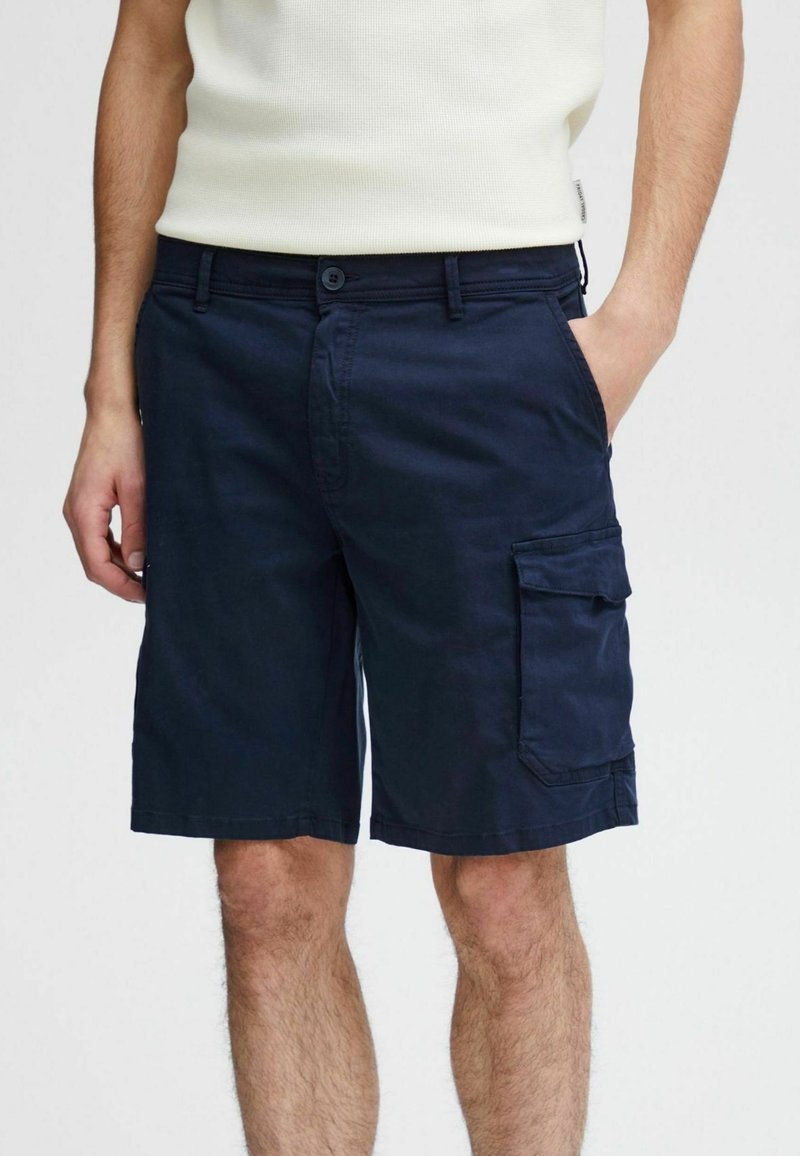 Homme portant un short cargo bleu foncé avec une poche à rabat latérale et un haut sans manches blanc, la main dans la poche, debout devant un fond uni.