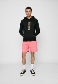 Svart hettegenser med grafikk av en tegneseriefigur, kombinert med rosa shorts. Modellen har på seg svarte sneakers og hvite sokker. Enkel bakgrunn.