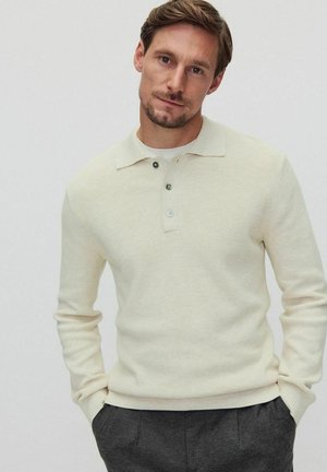 Vaaleanbeige neulepolo, jossa on pitkät hihat, kaksi vihreää nappia ja ribbikuvio. Sisältää klassisen kauluksen ja istuvan siluetin.