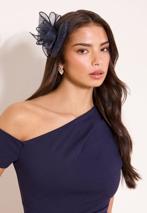 Femme aux longs cheveux bruns portant une robe bleu marine épaule dénudée et un grand accessoire floral en maille bleu marine sur le côté droit de sa tête.