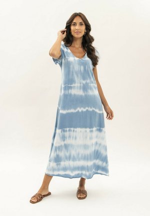 Femme portant une robe maxi tie-dye bleue et blanche à manches courtes et des sandales marron à enfiler, debout sur un fond blanc.