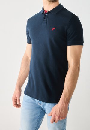 Homme portant un polo bleu marine à manches courtes avec un logo rouge brodé et un jean bleu clair, debout devant un fond uni clair.