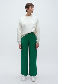 Λευκή φούτερ μπλούζα με χαλαρή εφαρμογή στους ώμους, συνδυασμένη με πράσινο παντελόνι φαρδιάς γραμμής. Το μοντέλο φοράει λευκά αθλητικά παπούτσια, παρουσιάζοντας μια άνετη εμφάνιση.