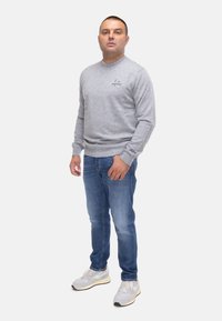 Felpa grigia con collo rotondo, tessuto testurizzato e piccolo ricamo. Indossata con jeans blu e scarpe da ginnastica grigie chiare.