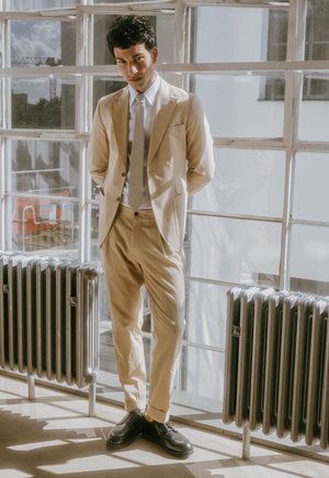 Man in beige suit and tie standing indoors by large window with metal radiators, hands behind back, looking forward.