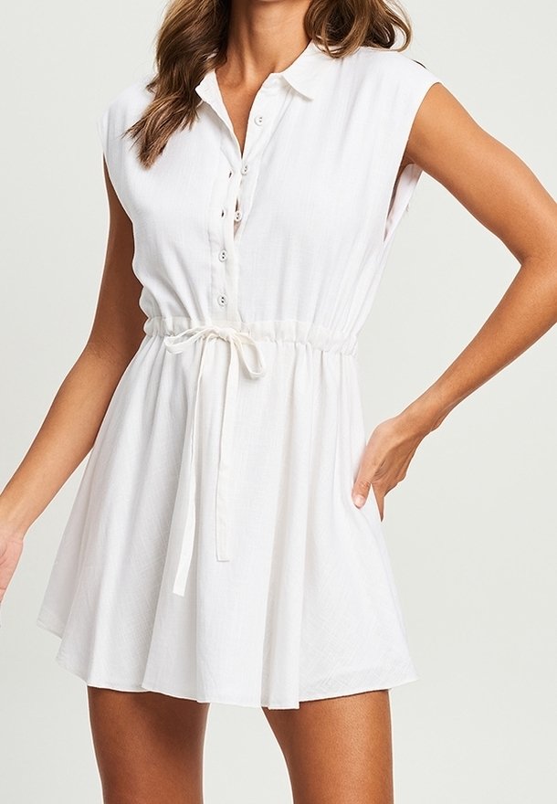 Femme portant une robe blanche sans manches avec des boutons sur le devant et une ceinture nouée à la taille, debout avec une main sur la hanche.