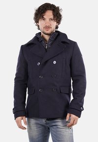 Cappotto navale doppiopetto con ampi revers, grandi bottoni e tasche laterali, indossato sopra una giacca interna trapuntata. Tessuto di lana liscia.