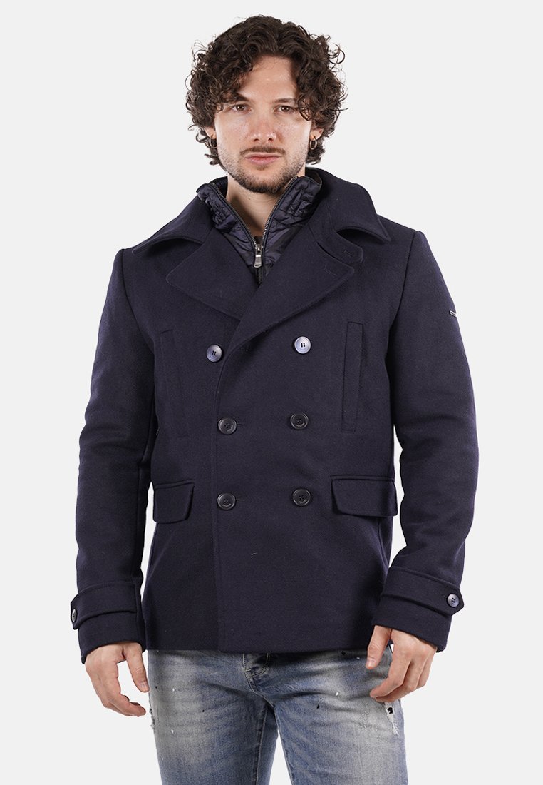 Cappotto navale doppiopetto con ampi revers, grandi bottoni e tasche laterali, indossato sopra una giacca interna trapuntata. Tessuto di lana liscia.