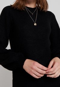 Pull en tricot noir avec des manches longues ajustées et un col rond. Colliers en or superposés avec des pendentifs ronds et en forme de feuille.