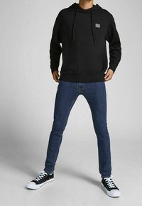 Person wearing black hoodie, blue skinny jeans, and black-and-white sneakers standing with legs apart on plain gray background.
