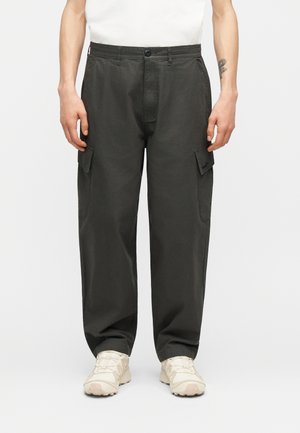 Mees, kes kannab tumedat halli vangapükse küljetaskutega ja valgeid tossusid, seisab tavalise valge tausta ees.